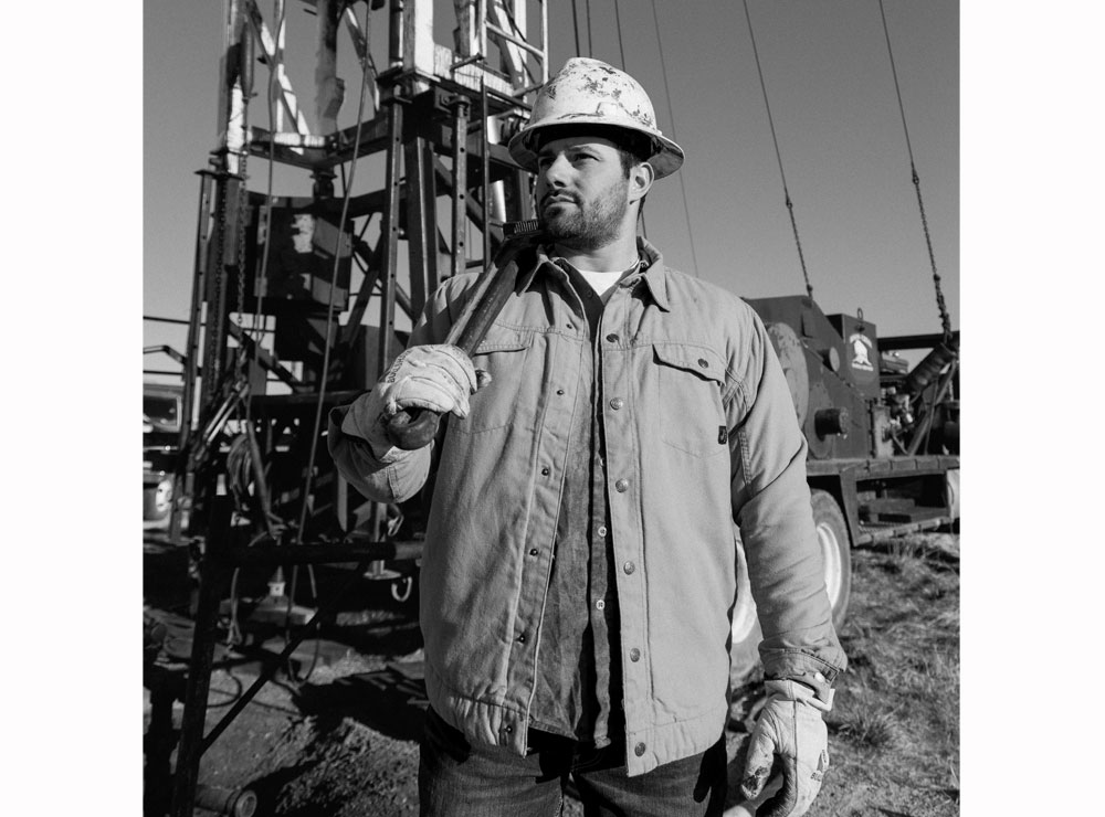Una fotografía en blanco y negro de un hombre sosteniendo una llave inglesa, posando frente a una plataforma petrolera.